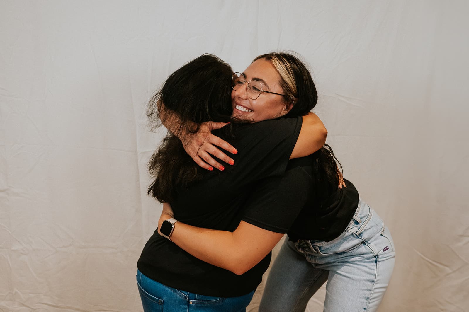 Nicole, wearing glasses and a black 'The Women's Wellbeing Club' t-shirt, sits against a softly lit wall, playfully running her hands through her hair. A warm-glowing floor lamp stands to her left, casting an inviting ambiance.
