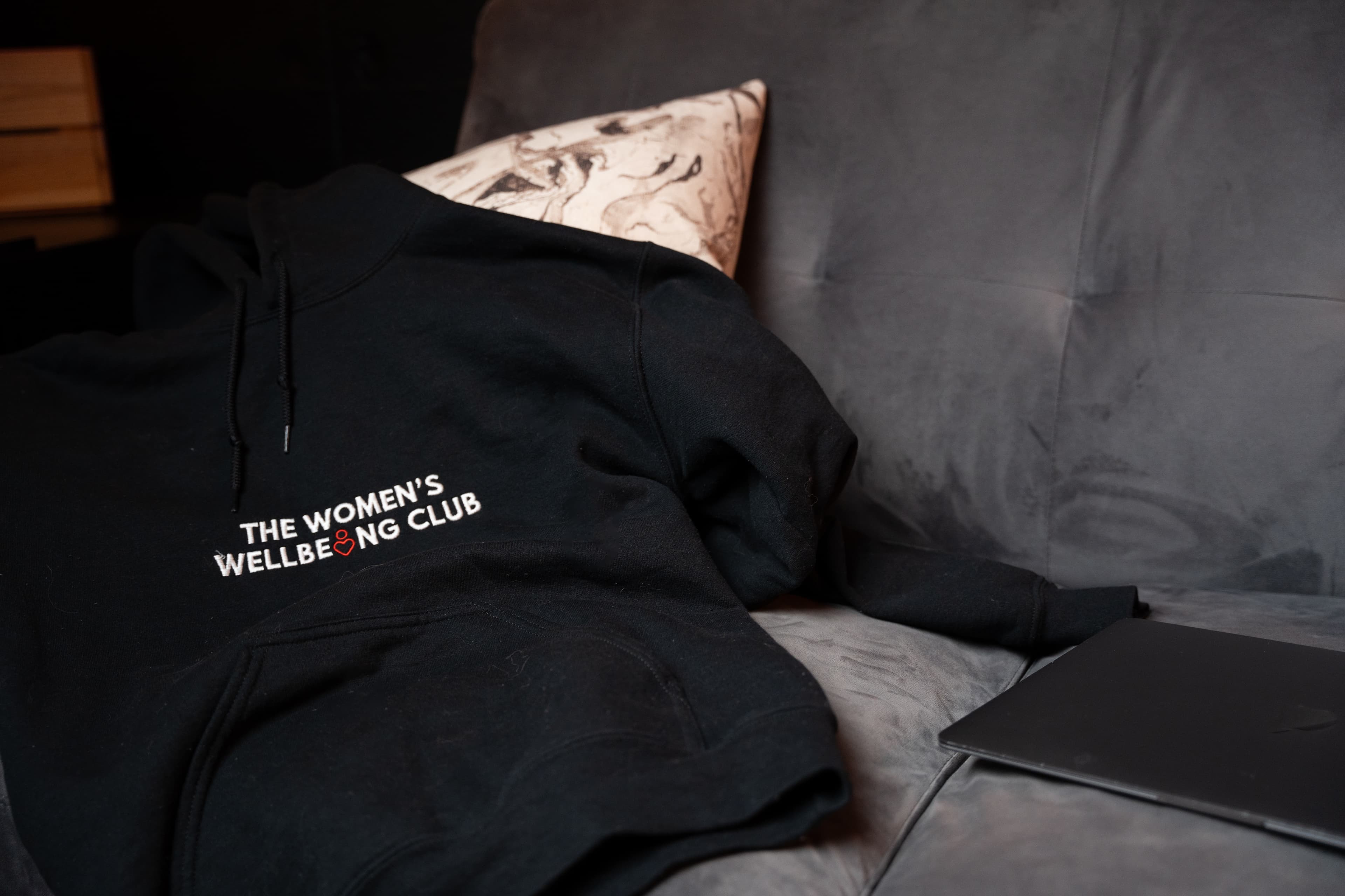 Nicole, wearing glasses and a black 'The Women's Wellbeing Club' t-shirt, sits against a softly lit wall, playfully running her hands through her hair. A warm-glowing floor lamp stands to her left, casting an inviting ambiance.