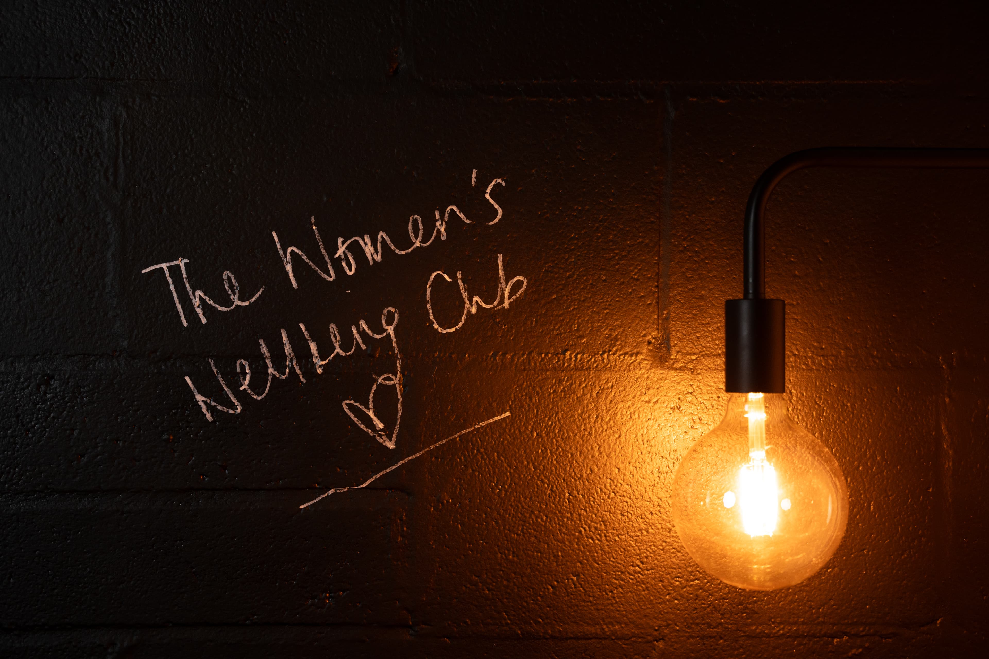 The Women's Wellbeing Club written on black wall with bulb lit up to the right.